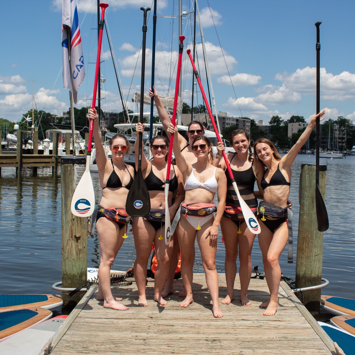 Bachelorette Party of 6 Ladies Standing on the Boardwalk with their Paddleboarding Gear Raised in the Air