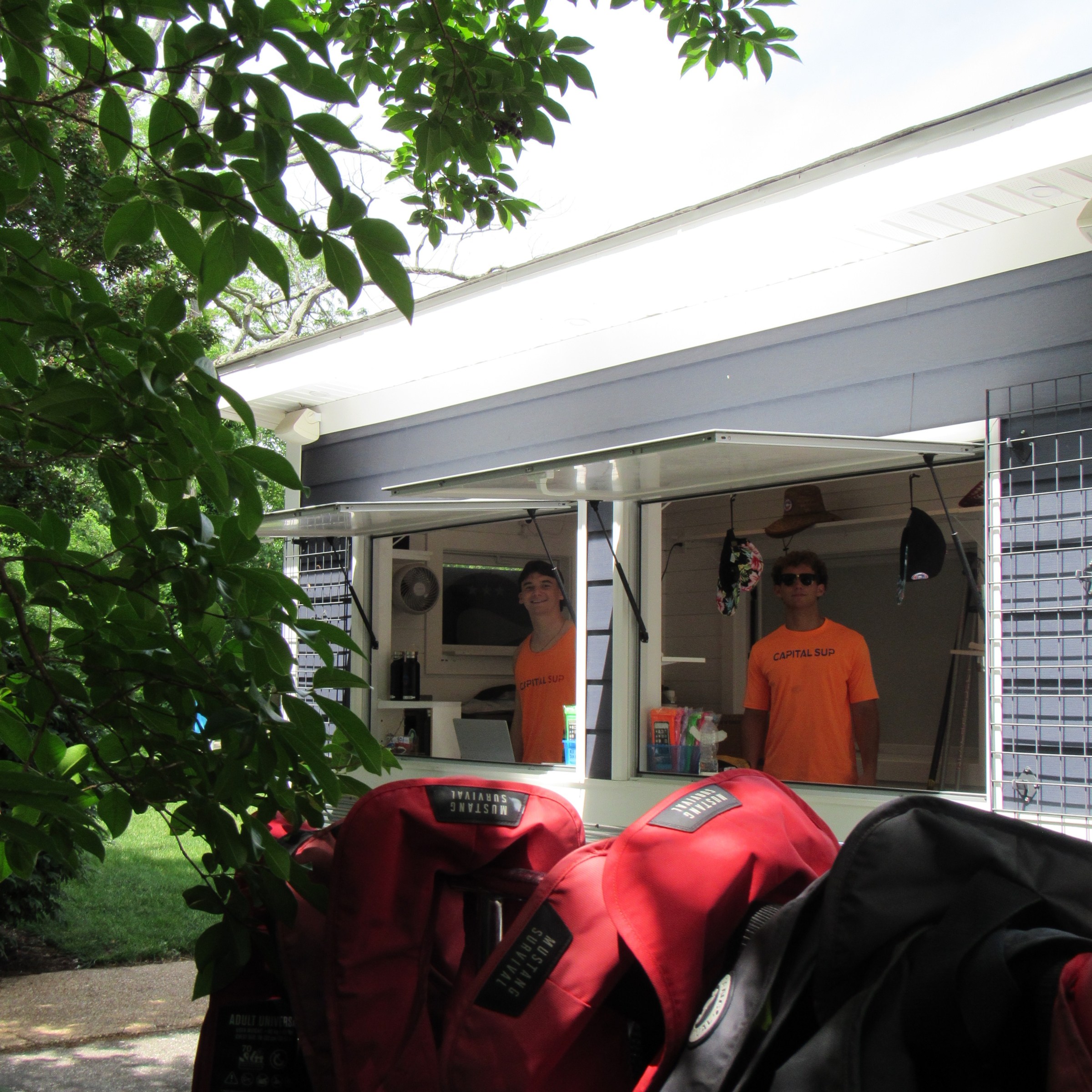 Two people in orange shirts stand in a kiosk with red life vests hanging in the foreground.