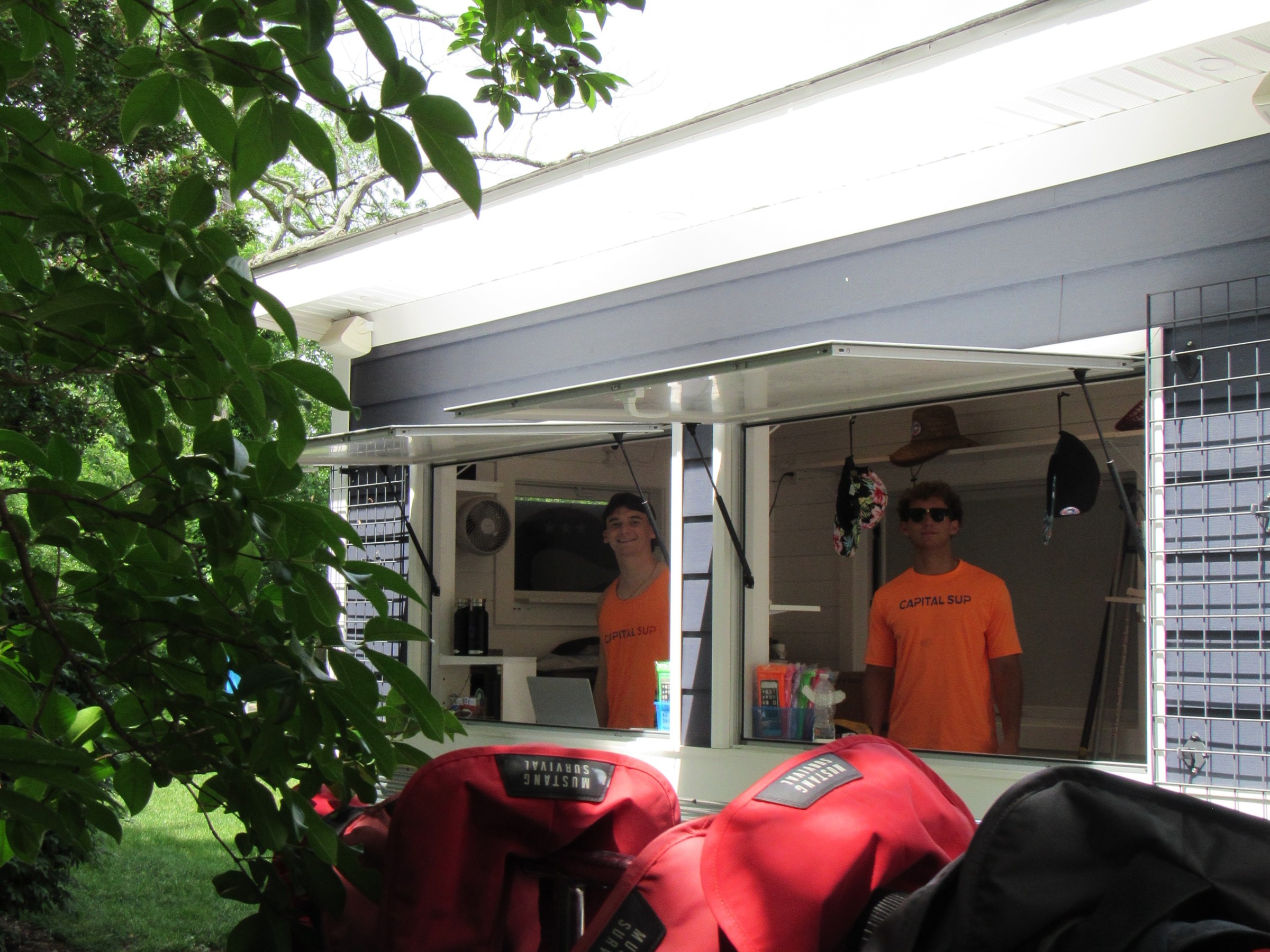 Two people in orange shirts stand in a kiosk with red life vests hanging in the foreground.