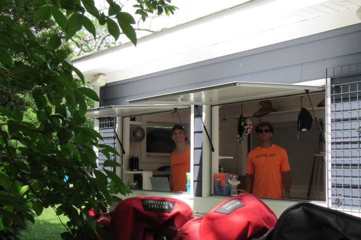 Two people in orange shirts stand in a kiosk with red life vests hanging in the foreground.