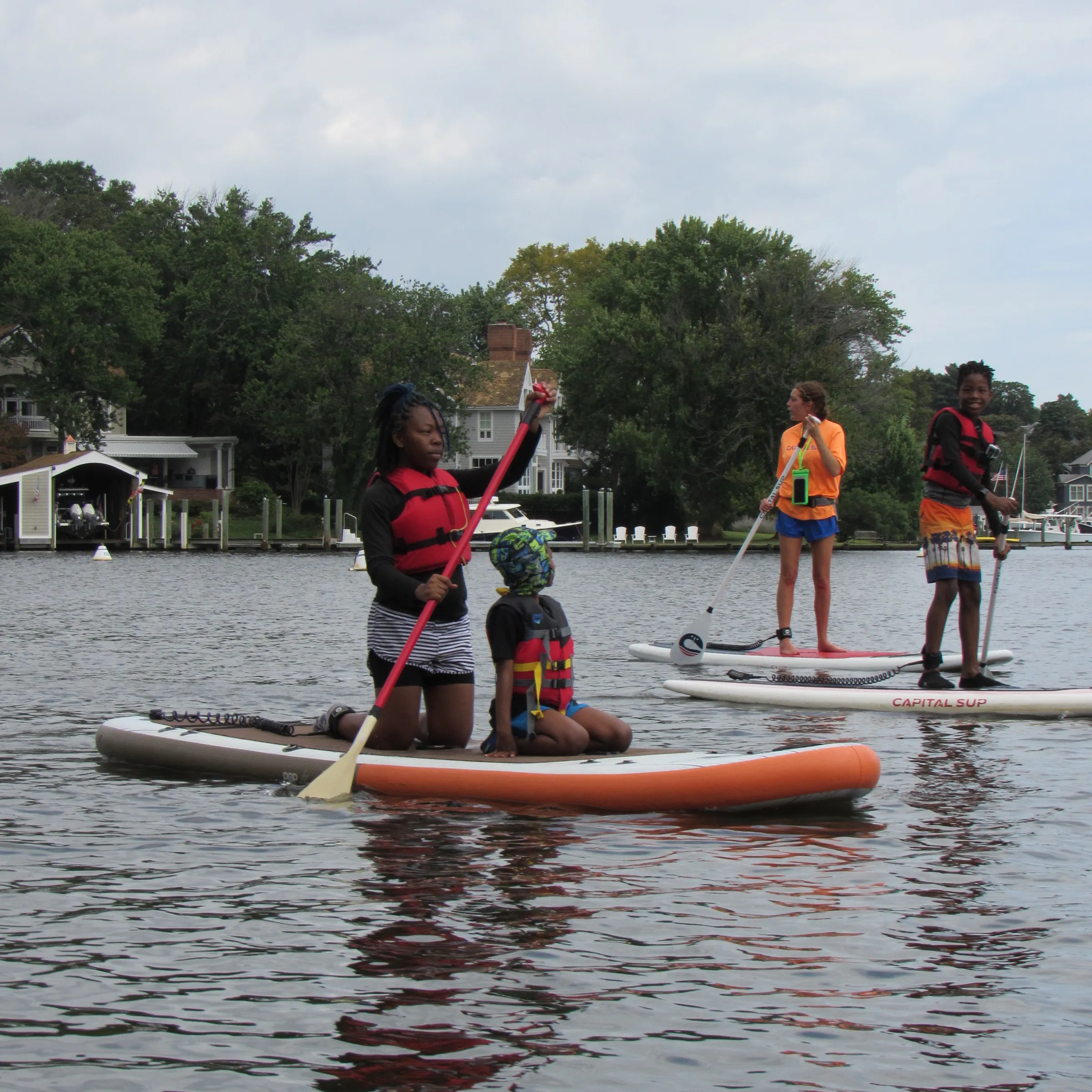 People paddleboarding on a lake with life vests and paddles, surrounded by trees and houses.