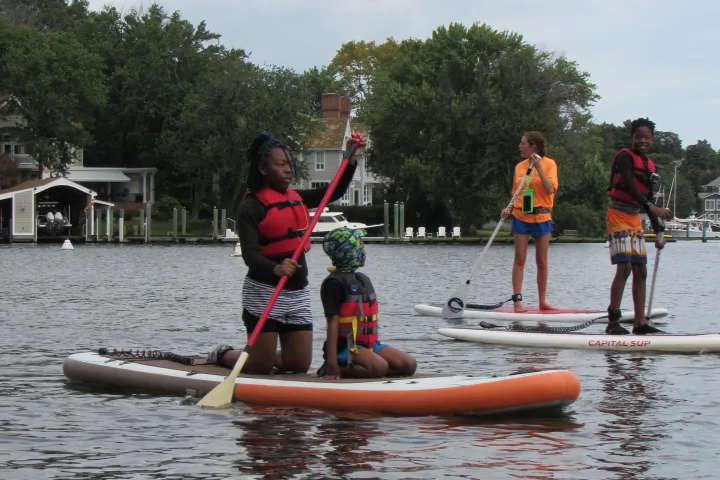People paddleboarding on a lake with life vests and paddles, surrounded by trees and houses.