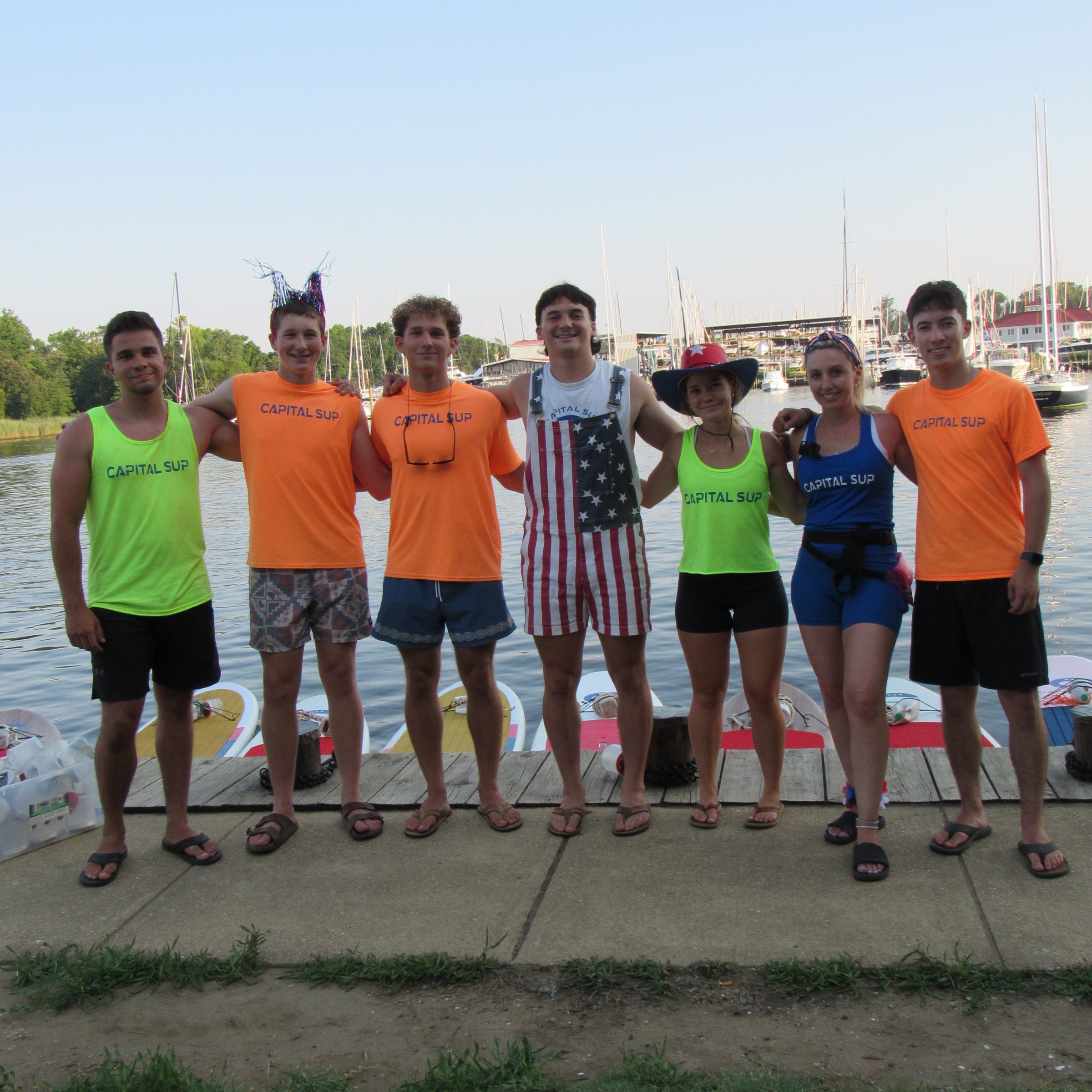 Group of seven people in colorful shirts by a waterfront, arms around each other, with paddleboards visible.