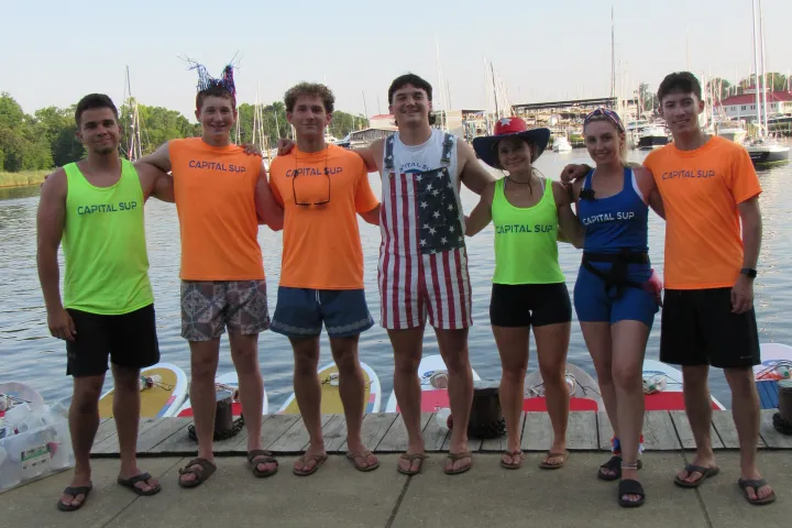 Group of seven people in colorful shirts by a waterfront, arms around each other, with paddleboards visible.