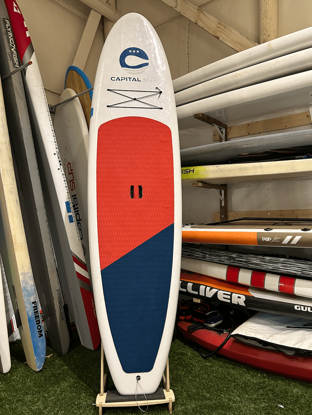 Screenshot 2025-08-08 at 2.38.20 PM Colorful paddleboard with 'Capital SUP' logo propped up in a board storage area.