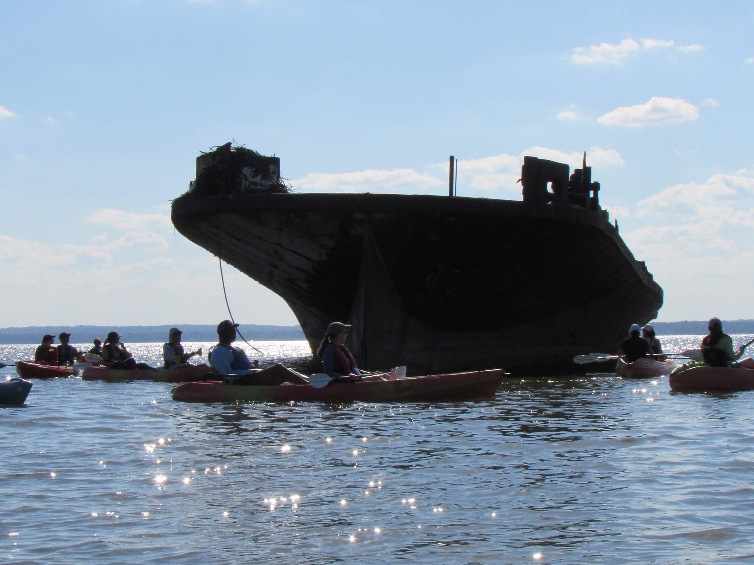 Kayakers surround a large, old shipwreck silhouette against a blue sky.