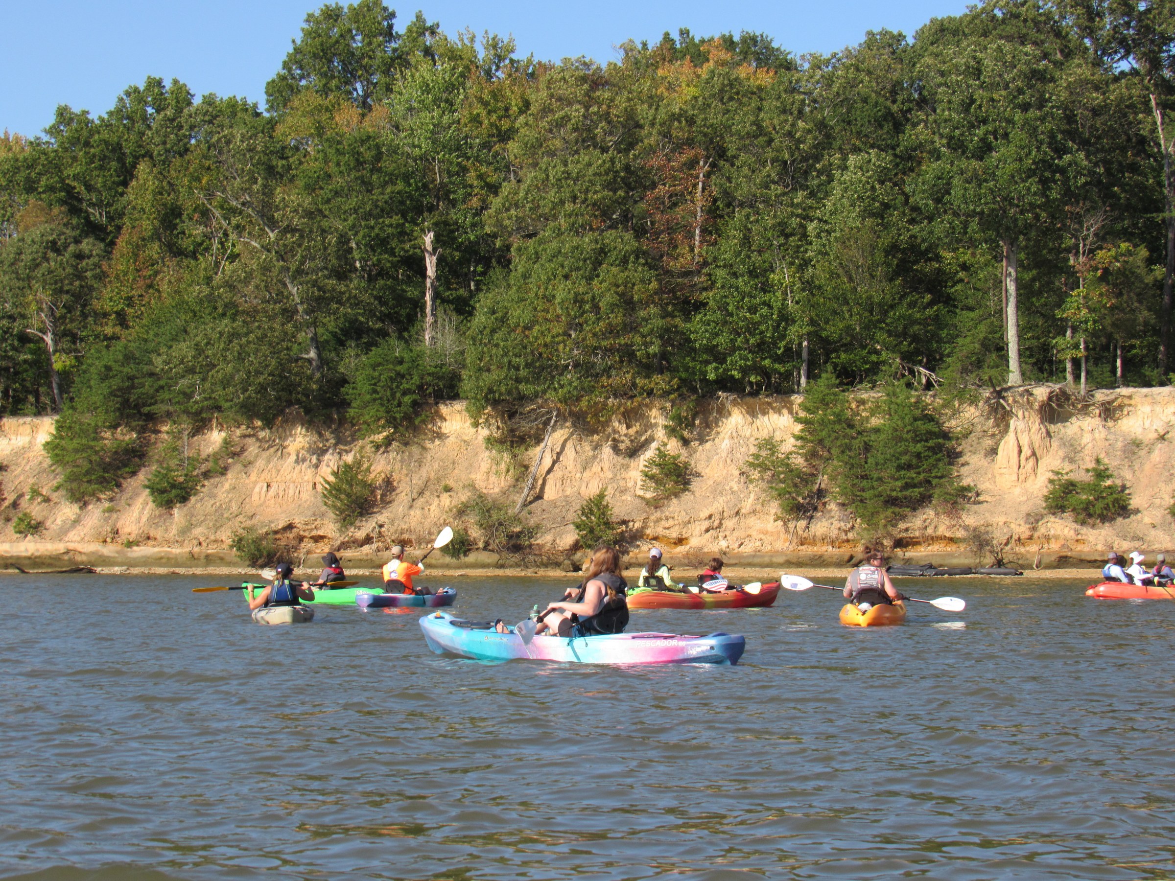 People kayaking on a lake near a forested shoreline under clear skies.