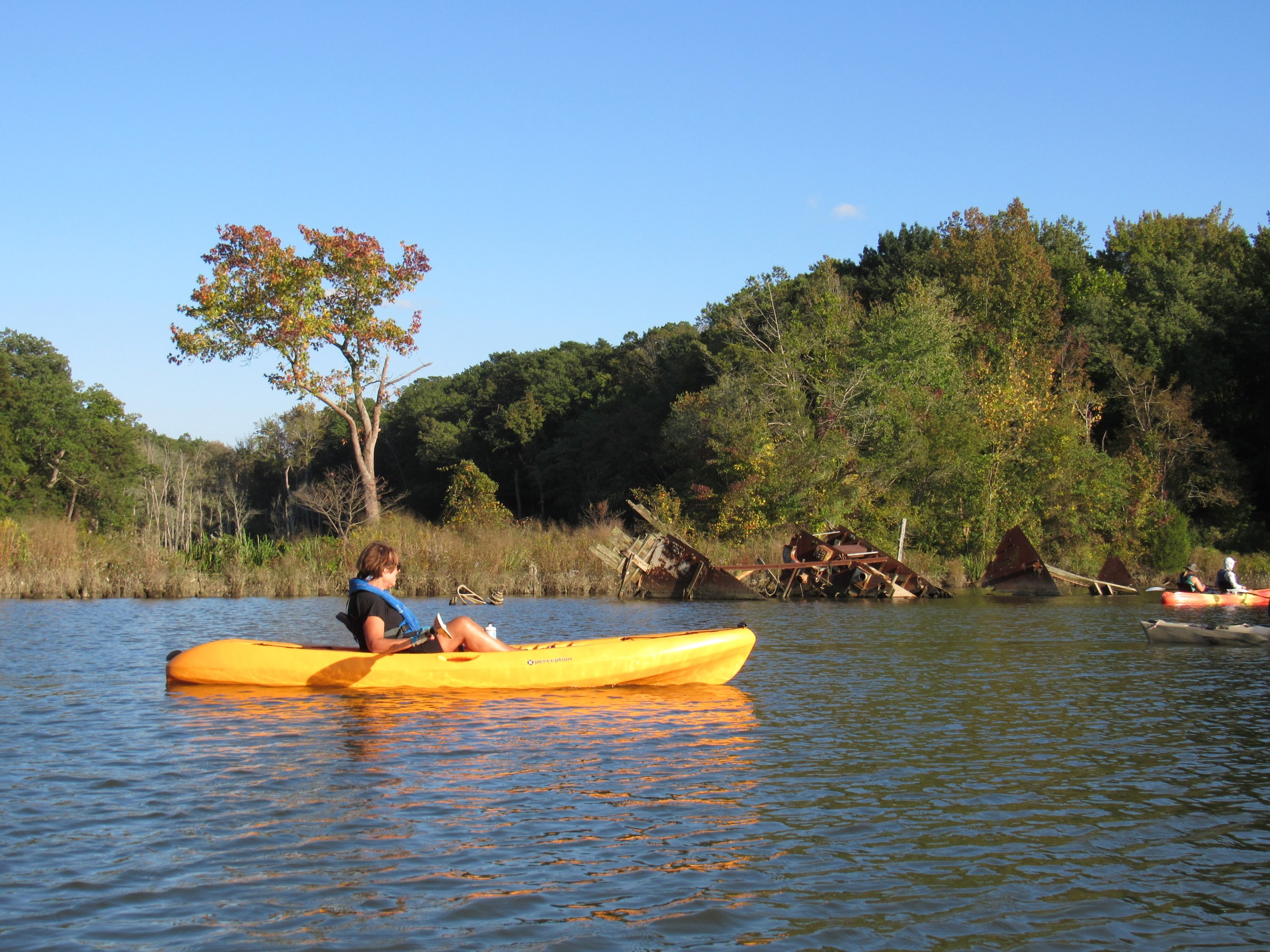 Person kayaking in a yellow kayak on a lake, with trees and a rusty shipwreck in the background.