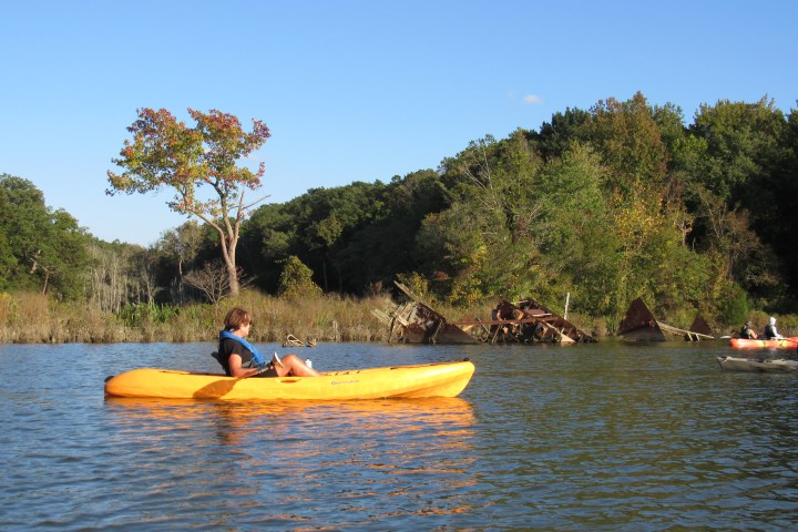 Person kayaking in a yellow kayak on a lake, with trees and a rusty shipwreck in the background.