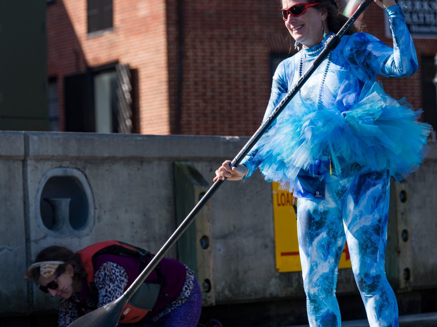 Person in blue costume and tutu paddleboarding near brick building.
