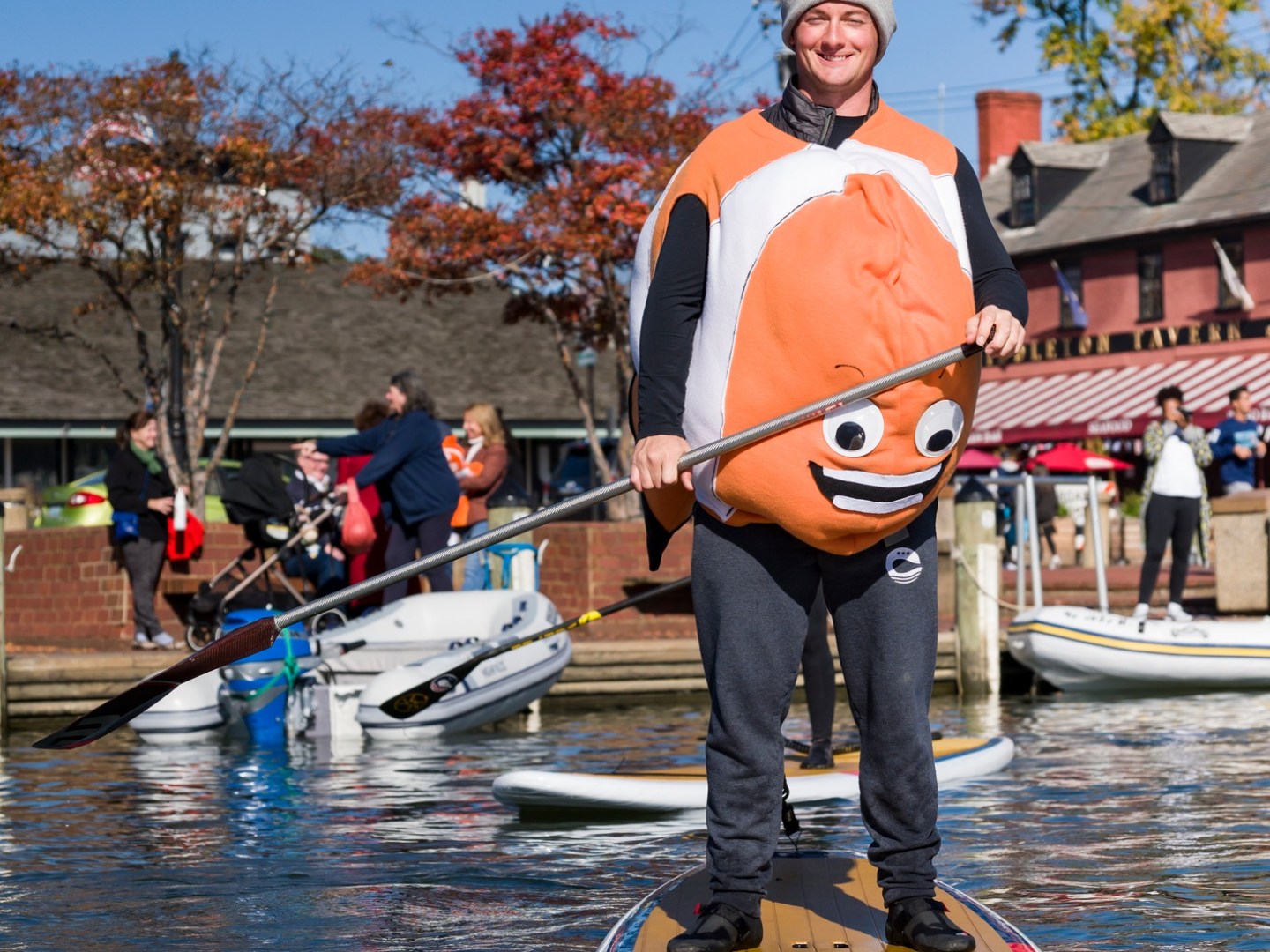 Person in a fish costume paddleboarding on a sunny day, with trees and people in the background.