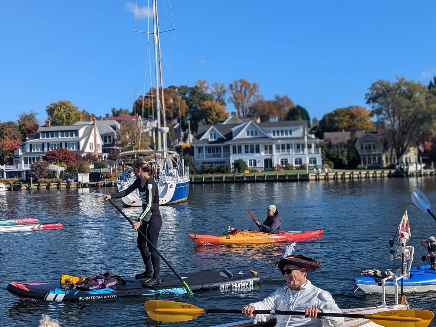 People in kayaks and paddleboards on a river with houses in background under a clear blue sky.