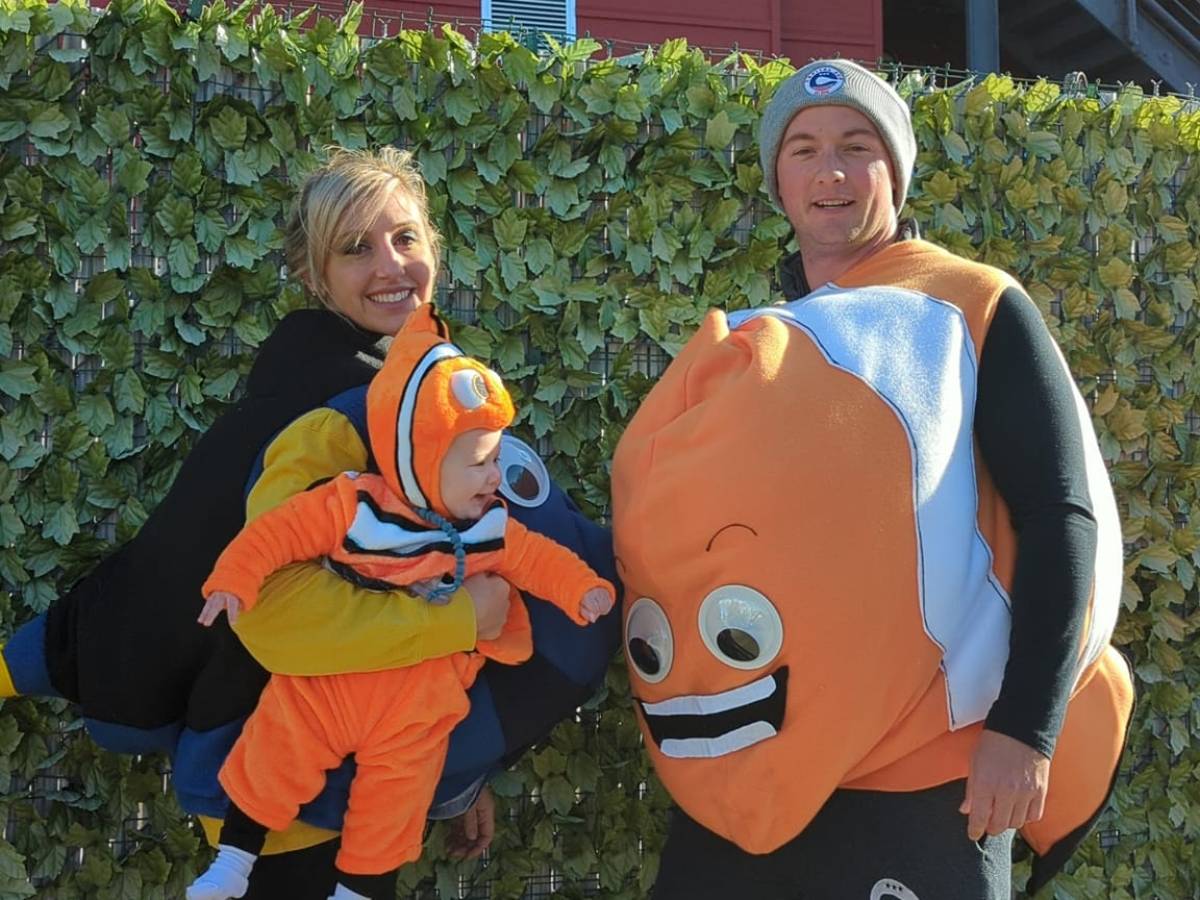 Three people dressed in cartoon fish costumes, standing in front of a hedge and red building.