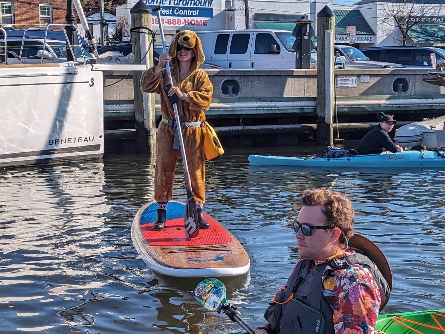 Person in bear costume paddleboarding near kayakers on a sunny day with boats and buildings in background.