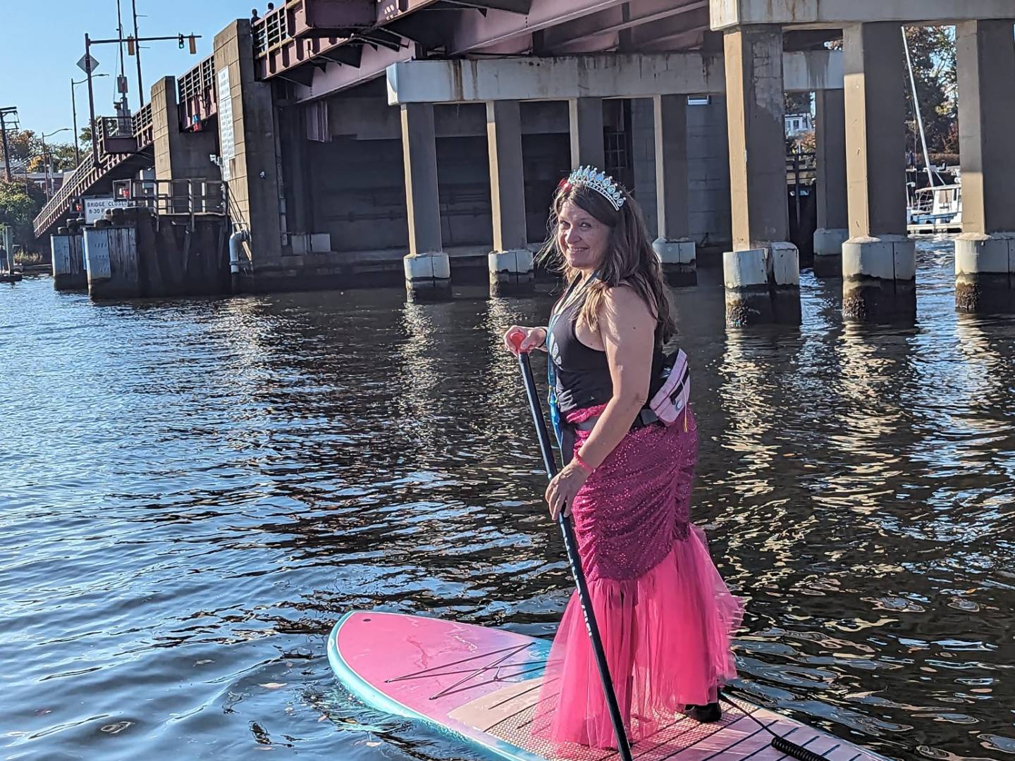 Woman in a pink dress and tiara paddleboarding under a bridge on a sunny day.
