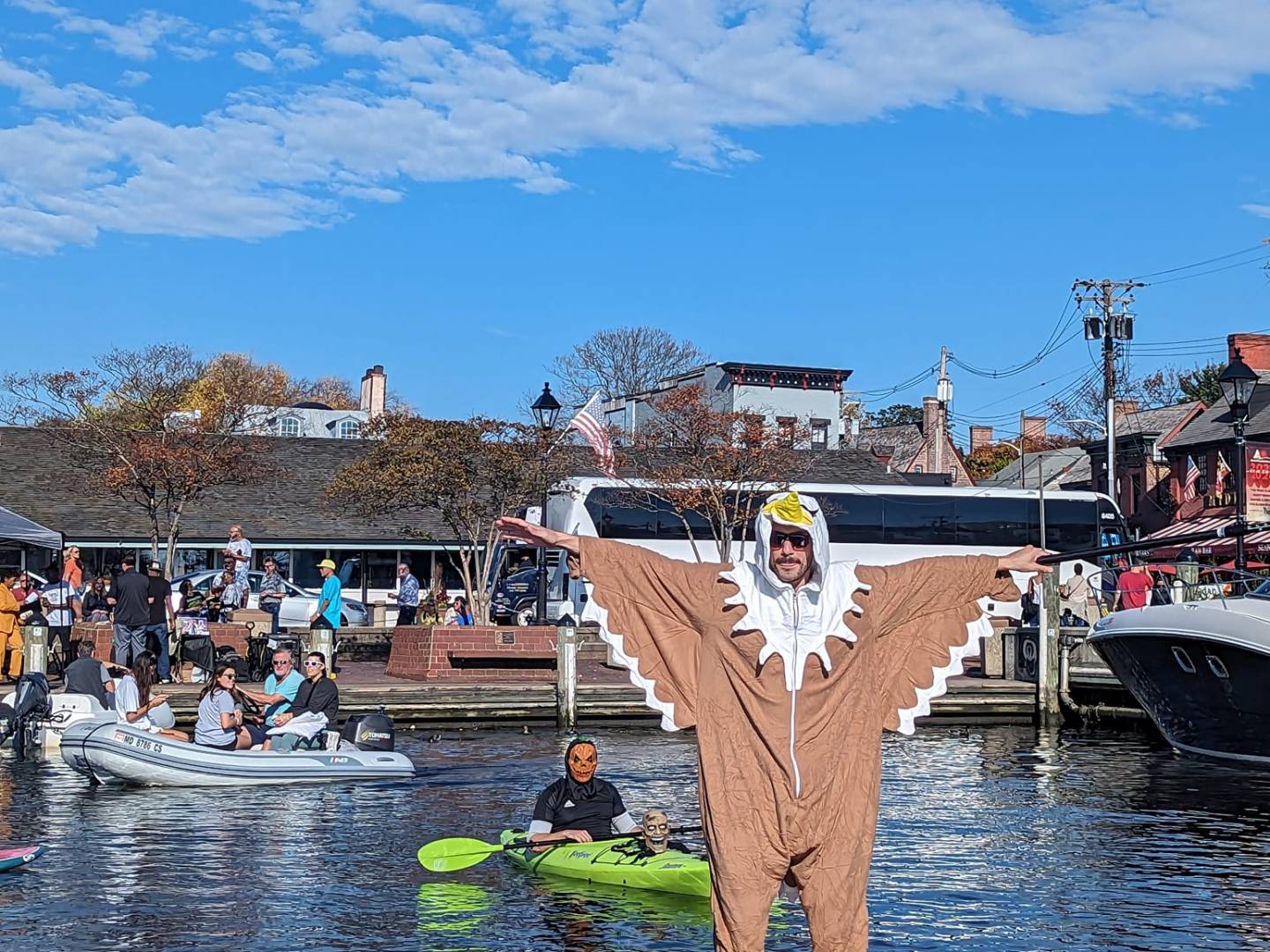Person in eagle costume paddleboarding on water, people in boats nearby, under clear blue sky.