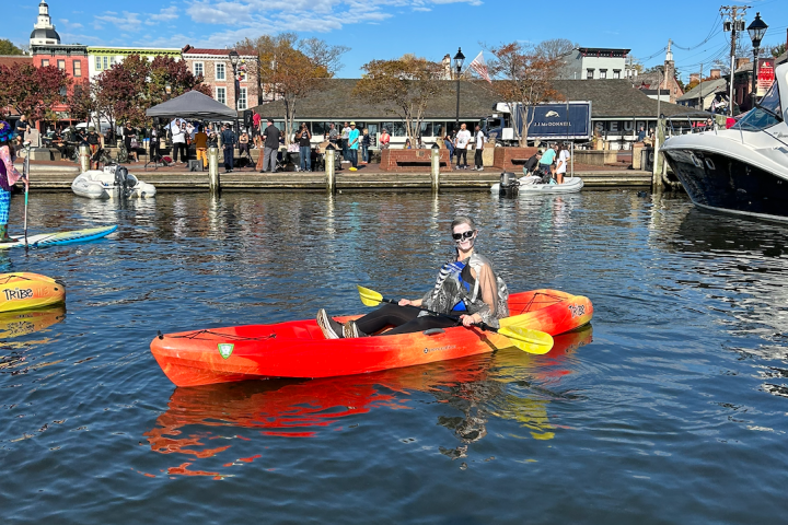Person in red kayak with yellow paddles on a sunny day, near crowded waterfront.
