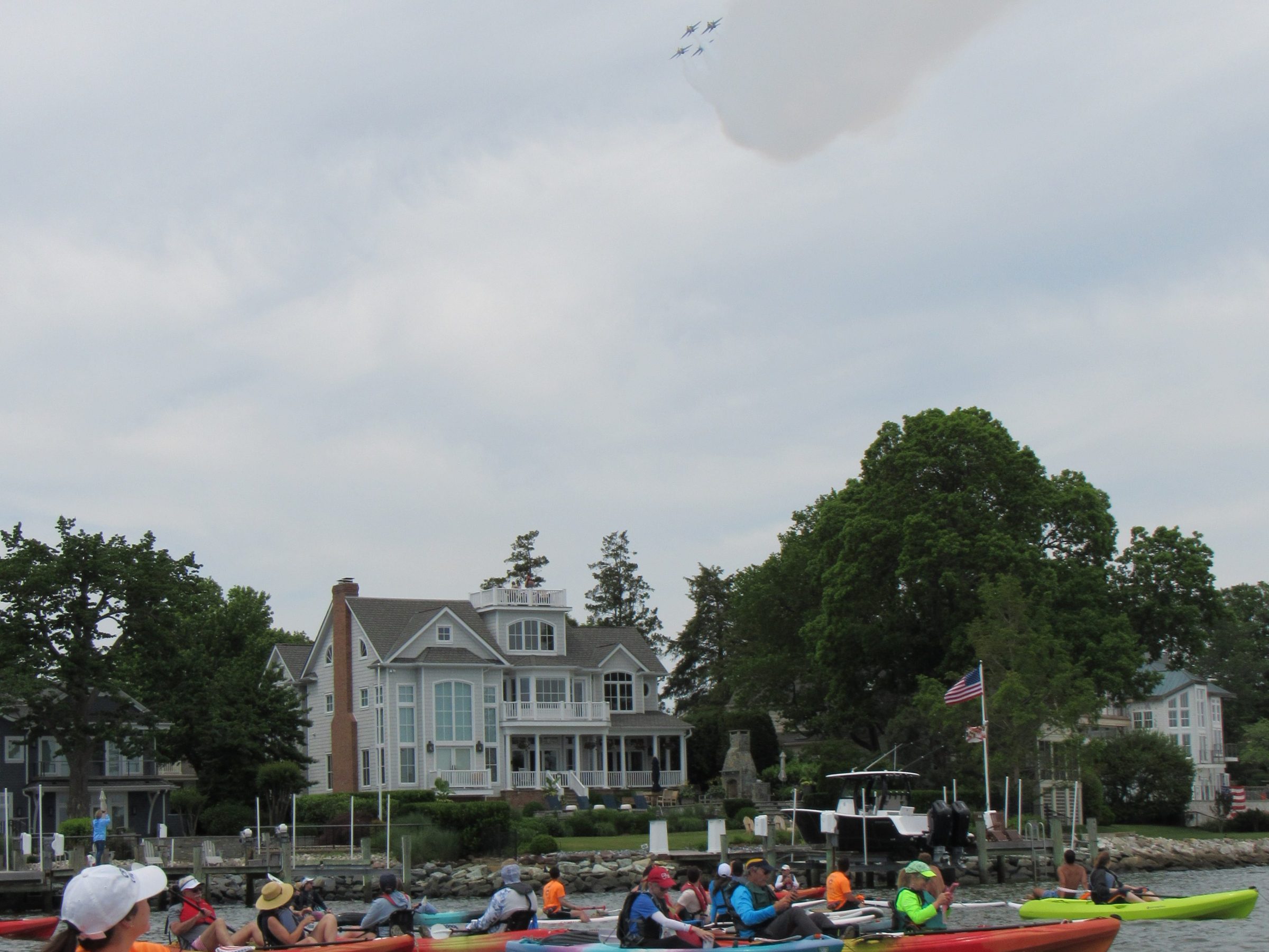 People kayaking on water, watching jets in the sky, with houses in the background.