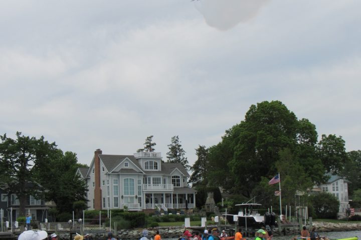People kayaking on water, watching jets in the sky, with houses in the background.