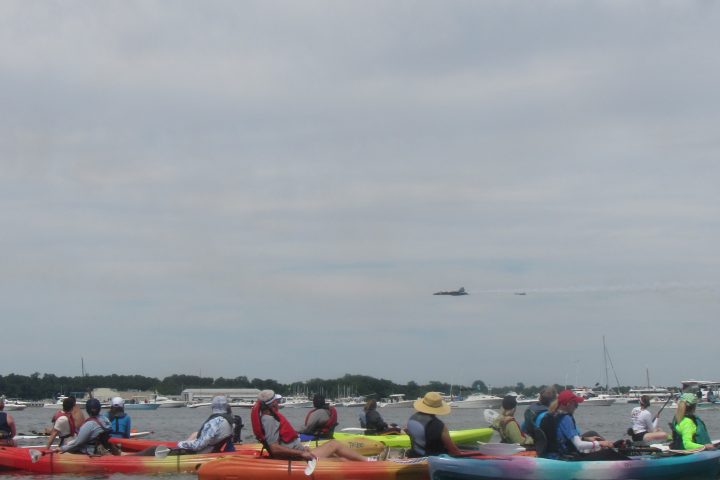 Kayakers on calm water watching a jet fly over during a cloudy day.