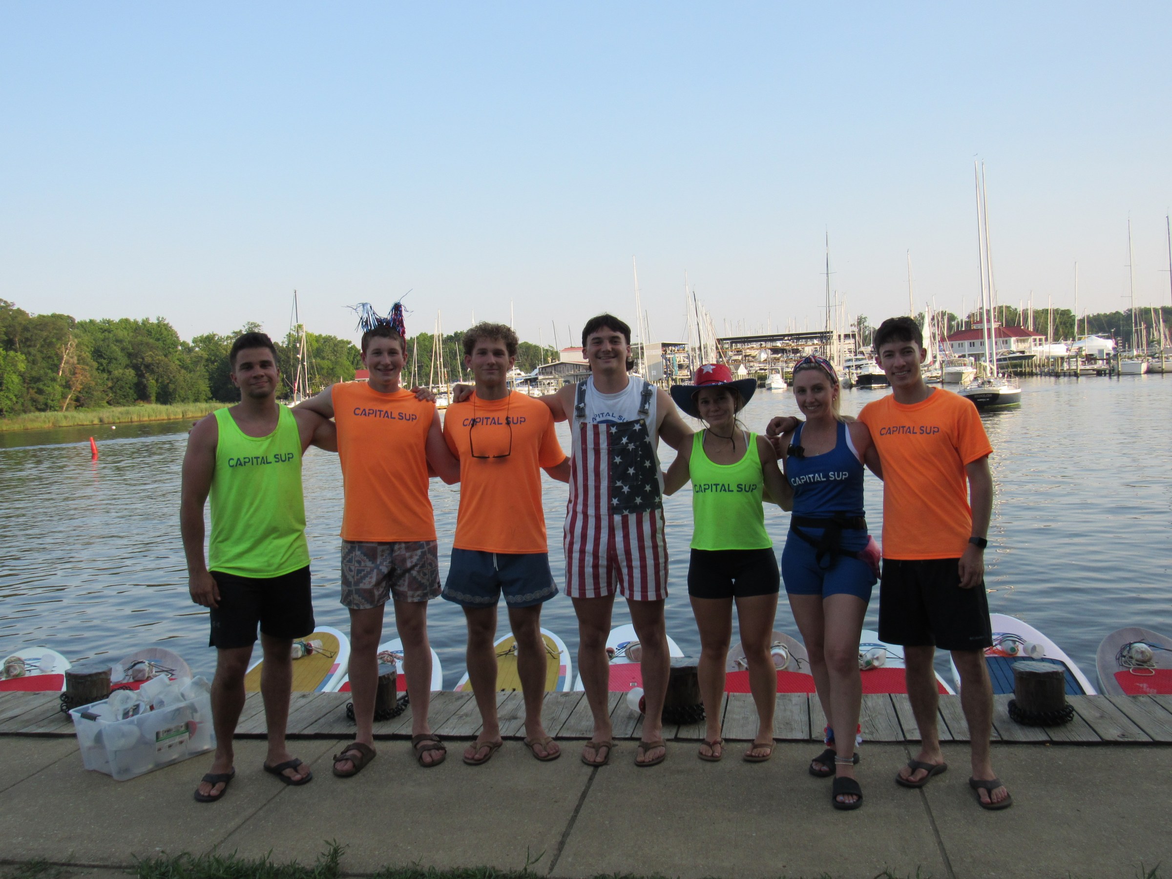 Group of seven people wearing Capital SUP shirts standing by a dock with boats in the background.