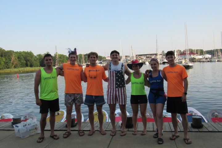 Group of seven people wearing Capital SUP shirts standing by a dock with boats in the background.