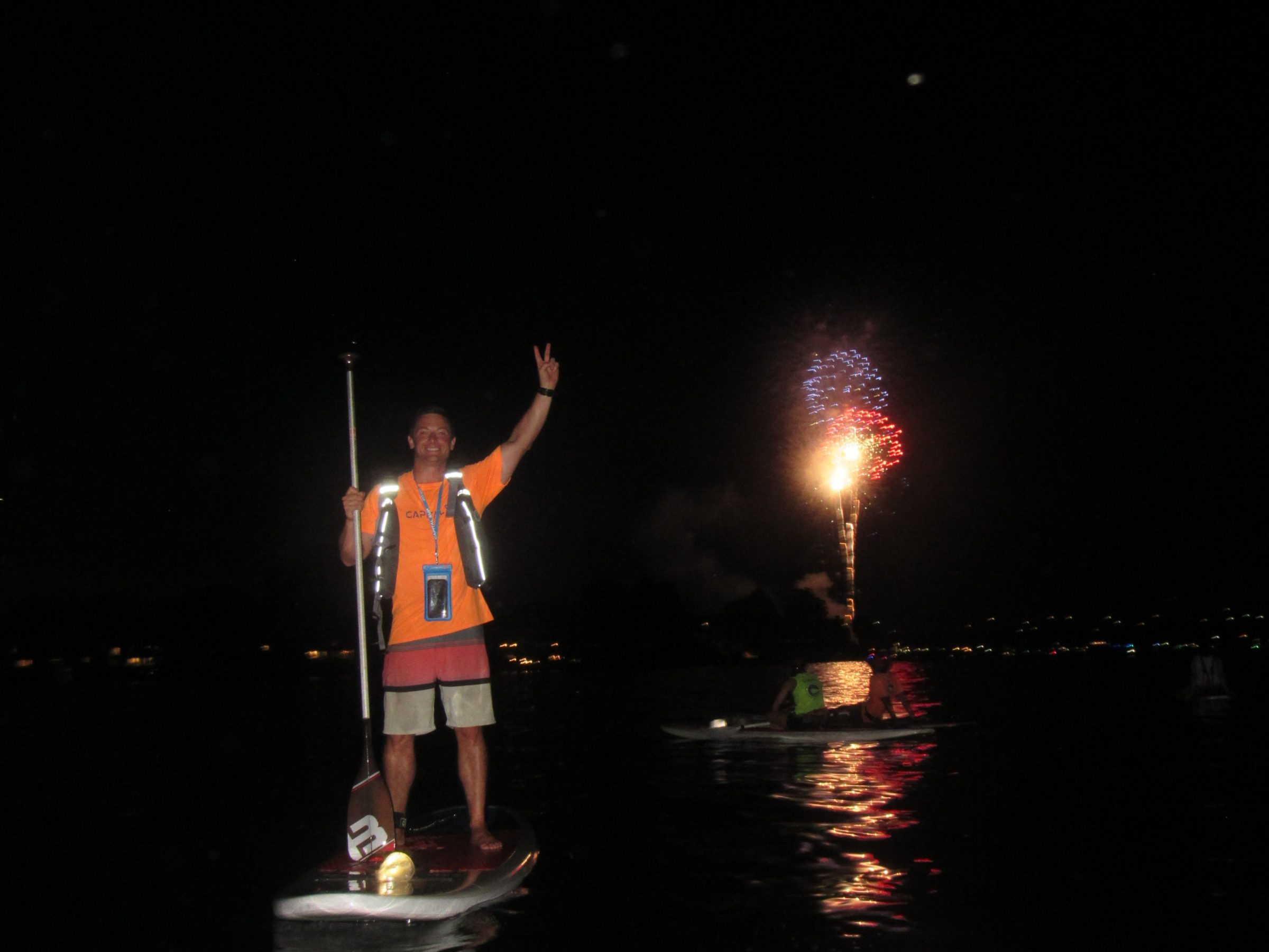 Person on paddleboard at night raising hand with fireworks in the background.