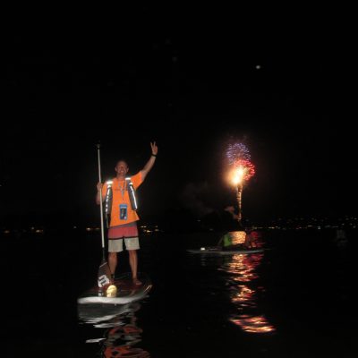 Person on paddleboard at night raising hand with fireworks in the background.