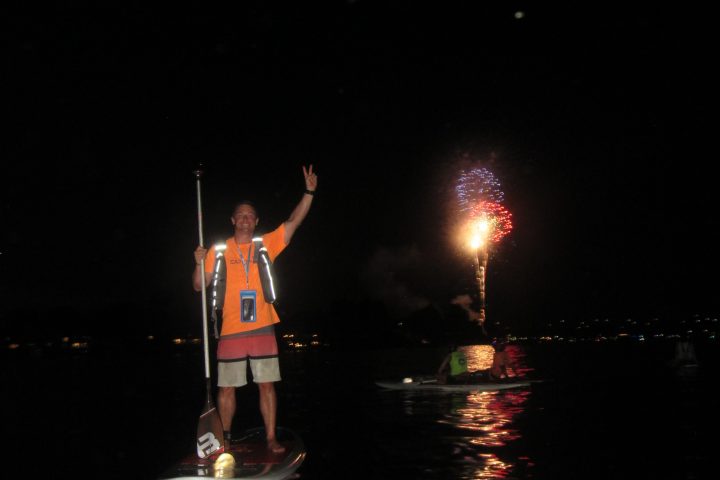 Person on paddleboard at night raising hand with fireworks in the background.