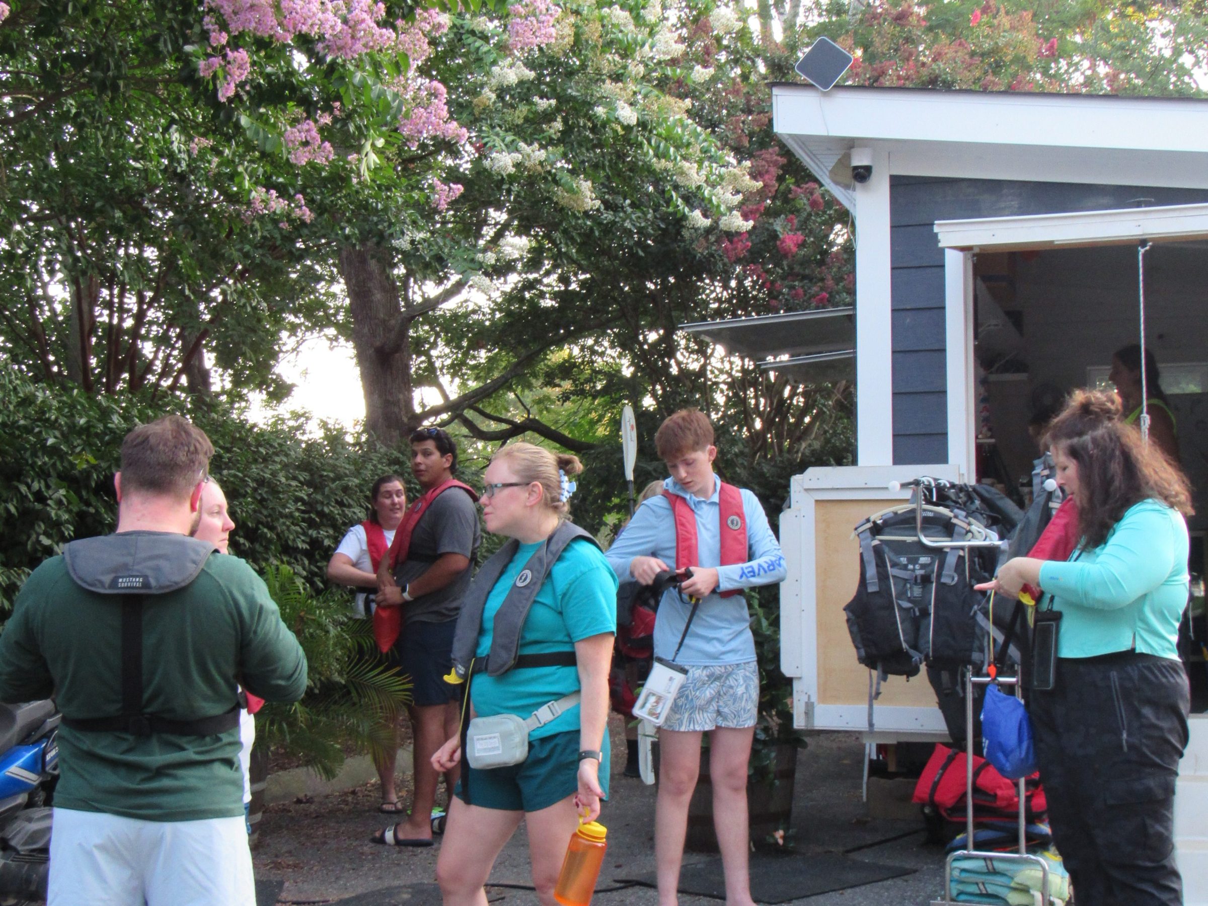 Group of people in life jackets preparing for outdoor activity near a house and flowering trees.