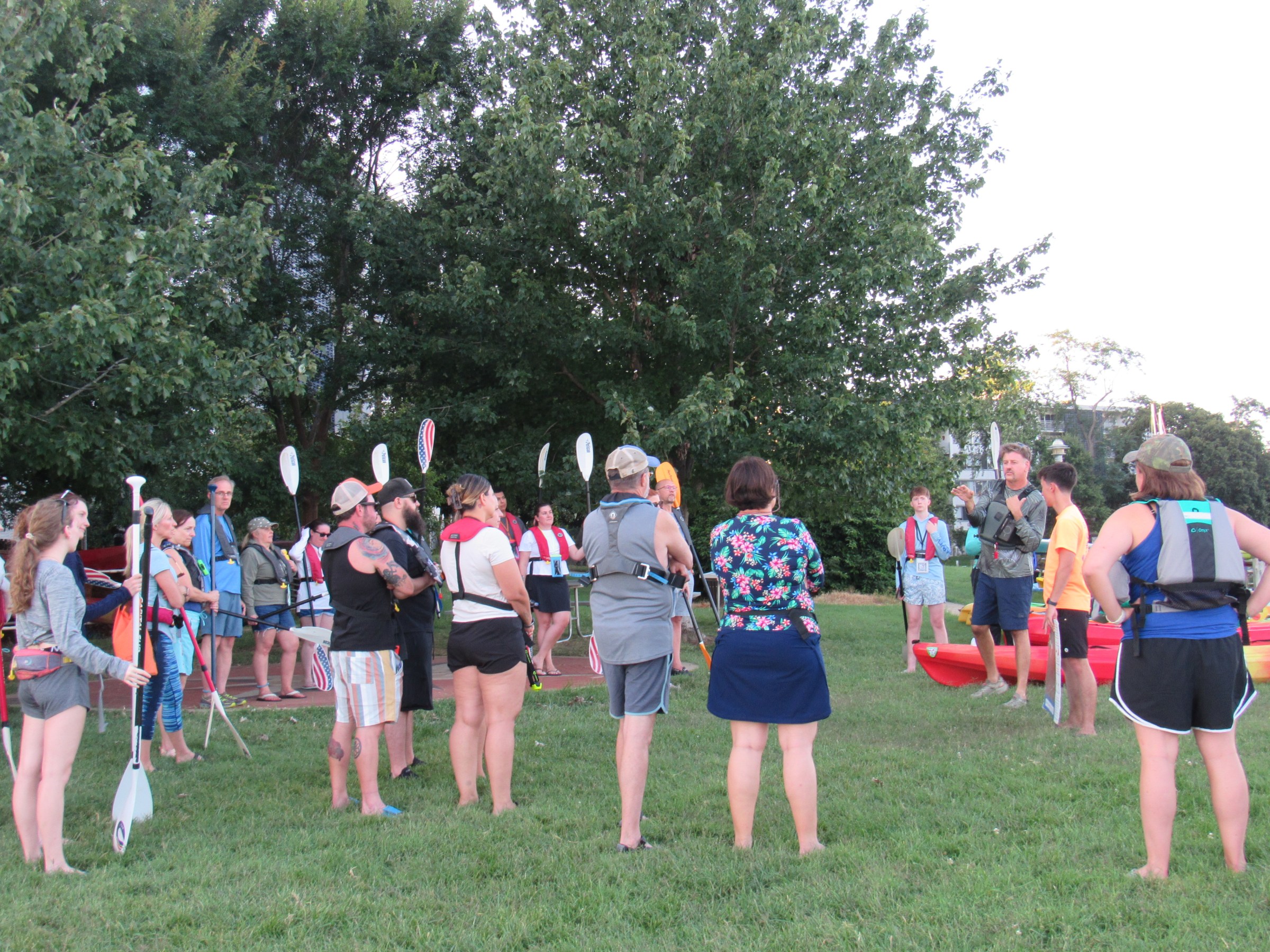 Group of people with paddles and life jackets gather on grass near kayaks.