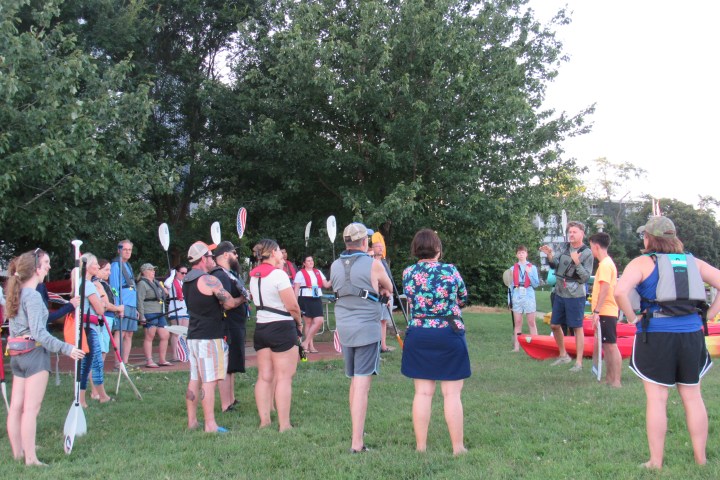Group of people with paddles and life jackets gather on grass near kayaks.