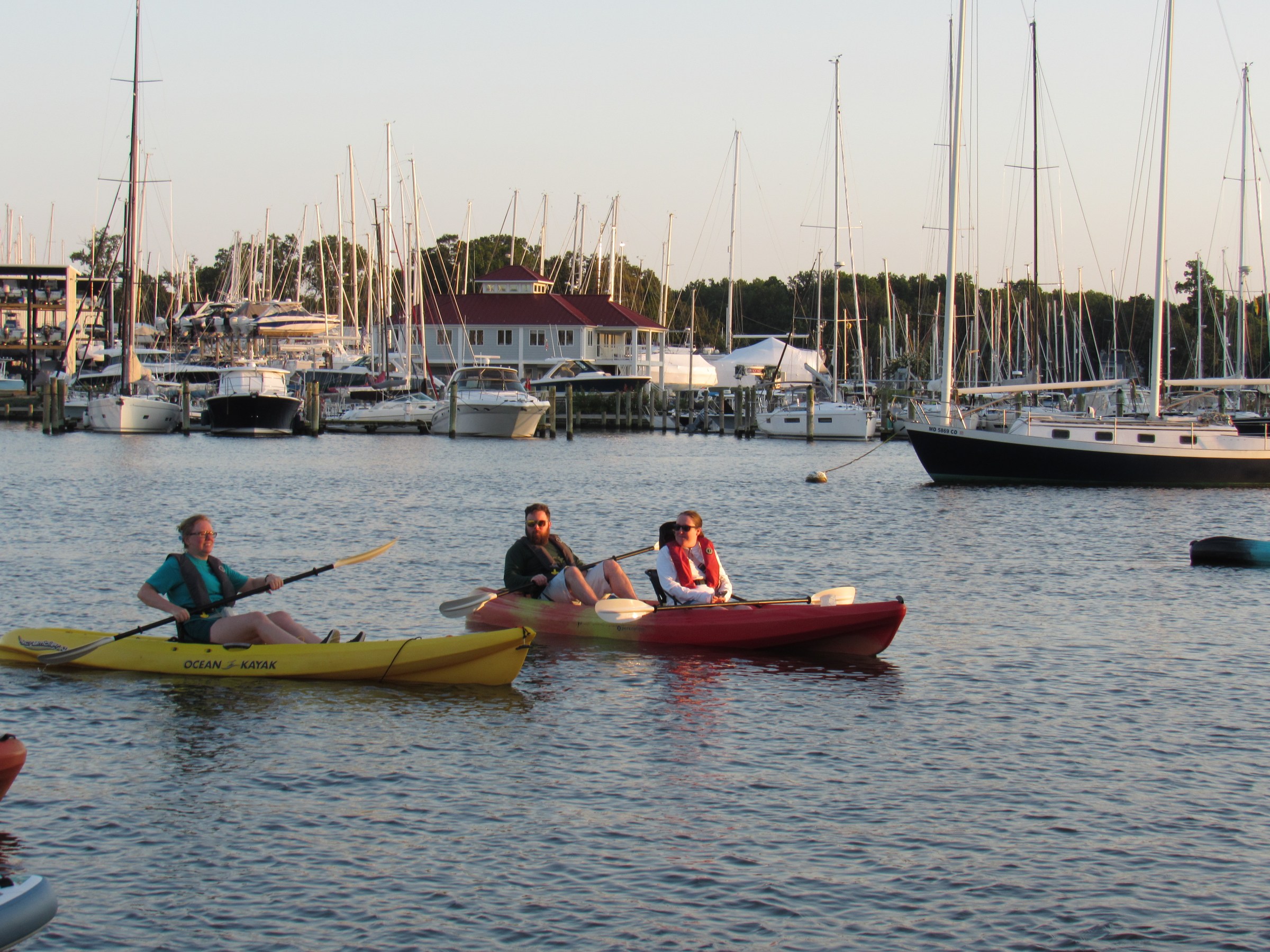 Three people kayaking near a marina with sailboats at sunset.