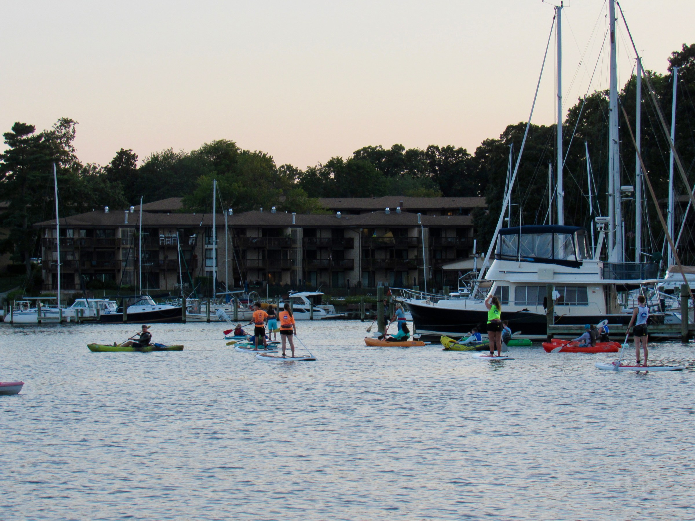 People kayaking and paddleboarding near docked boats at sunset.