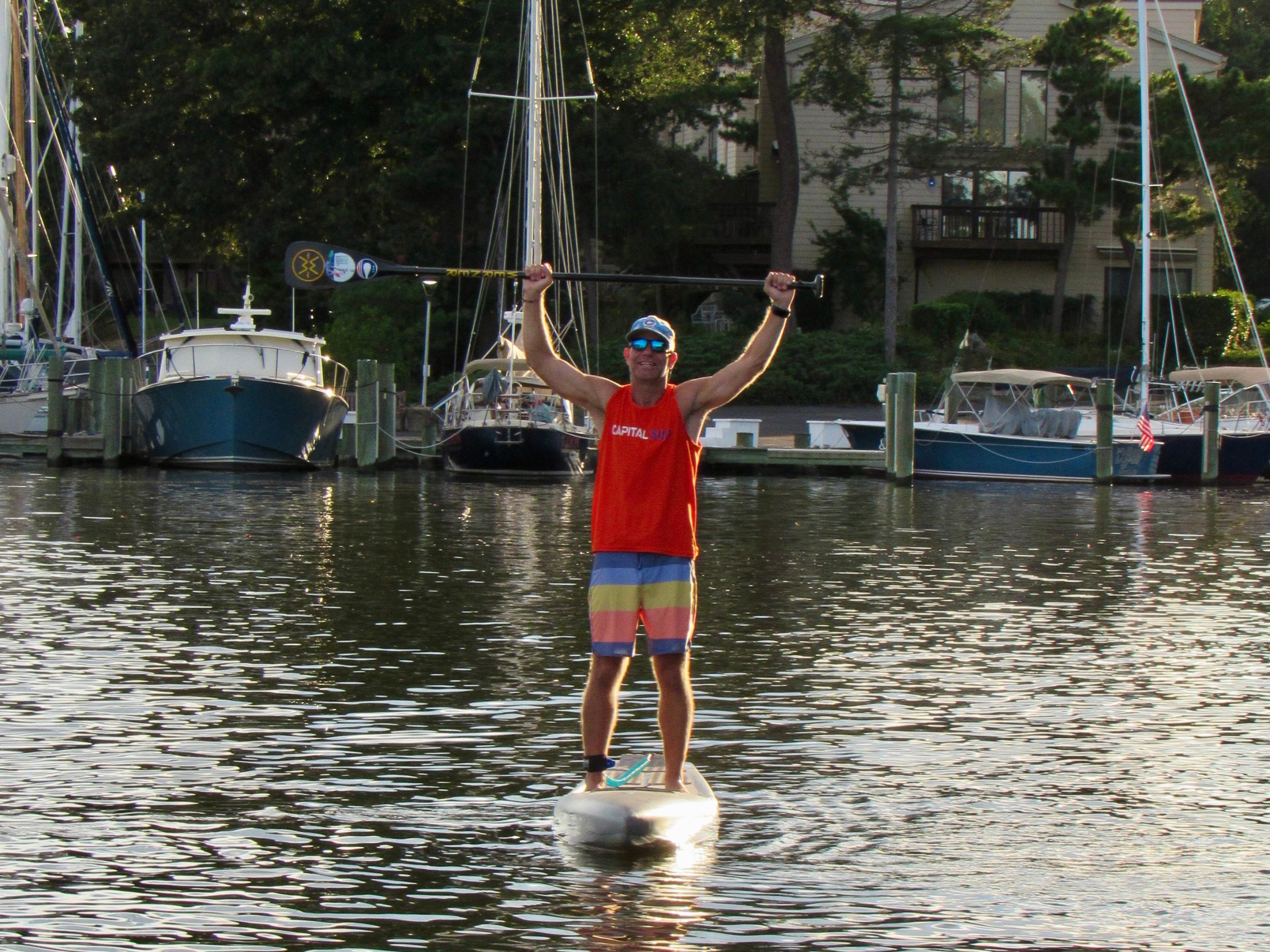 Person in red tank top paddleboarding on a calm river near docked sailboats.