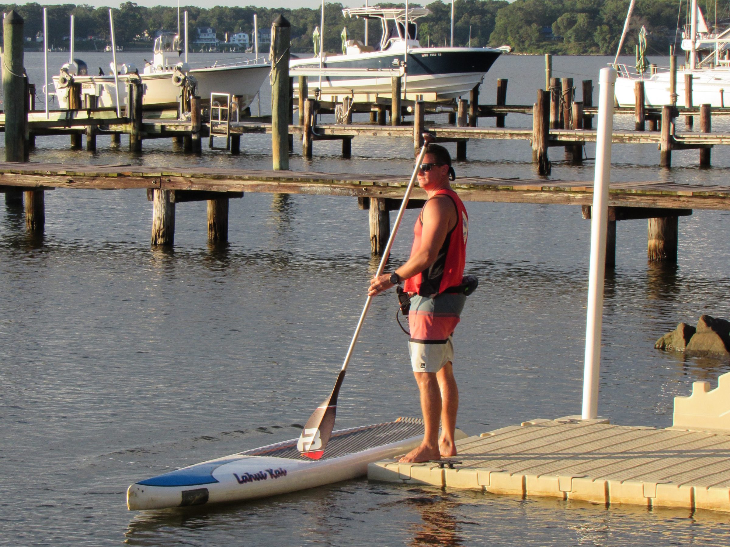 Person with paddleboard on dock next to calm water and boats in background.