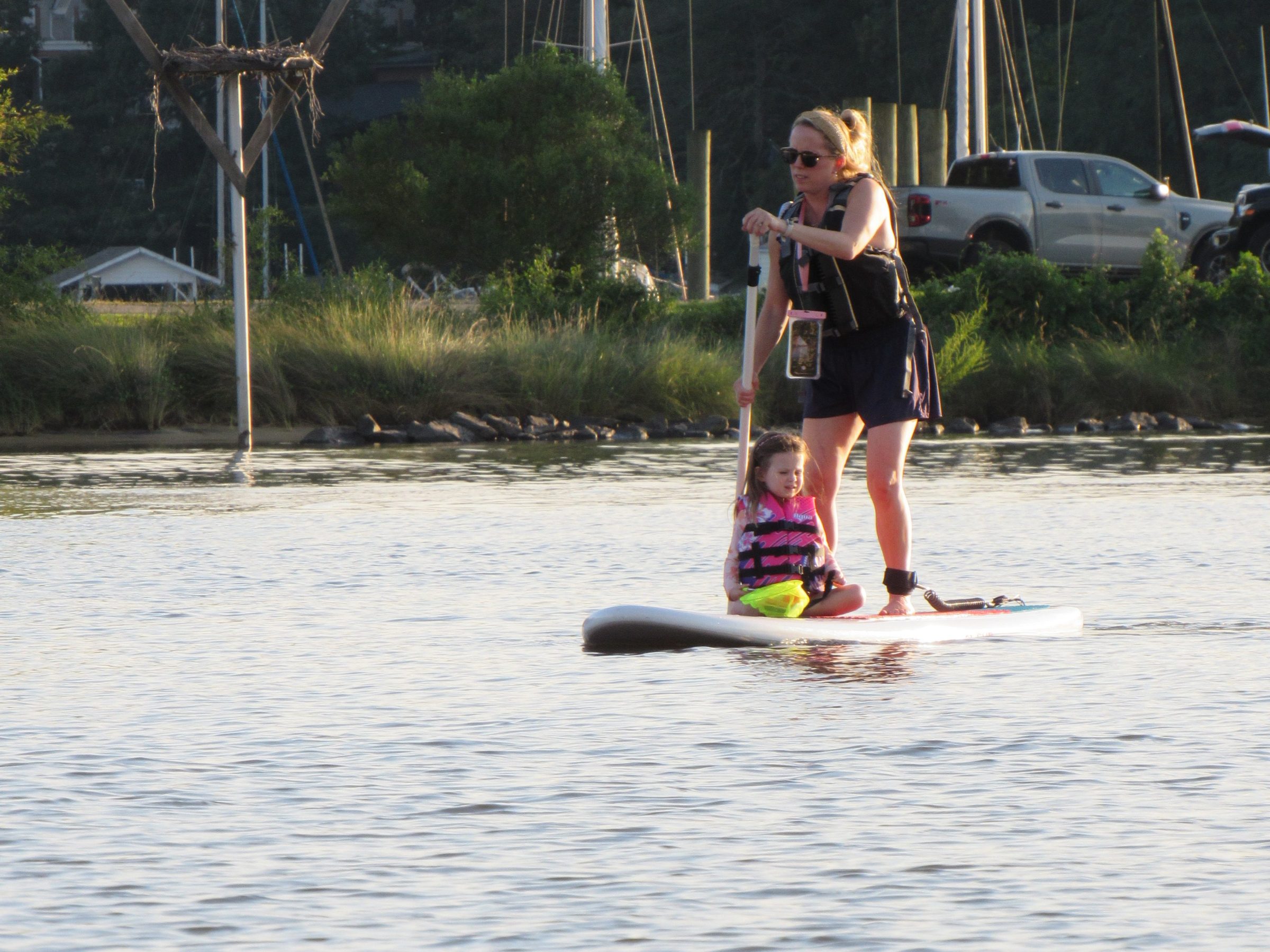 Woman paddleboarding with a child on a calm river, trees and boats in the background.