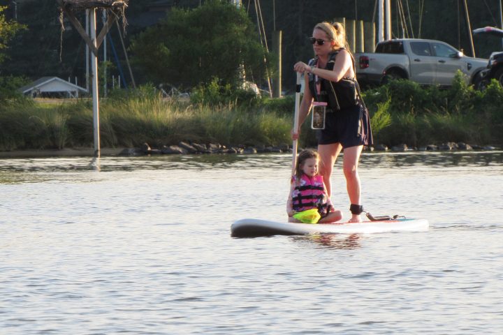 Woman paddleboarding with a child on a calm river, trees and boats in the background.