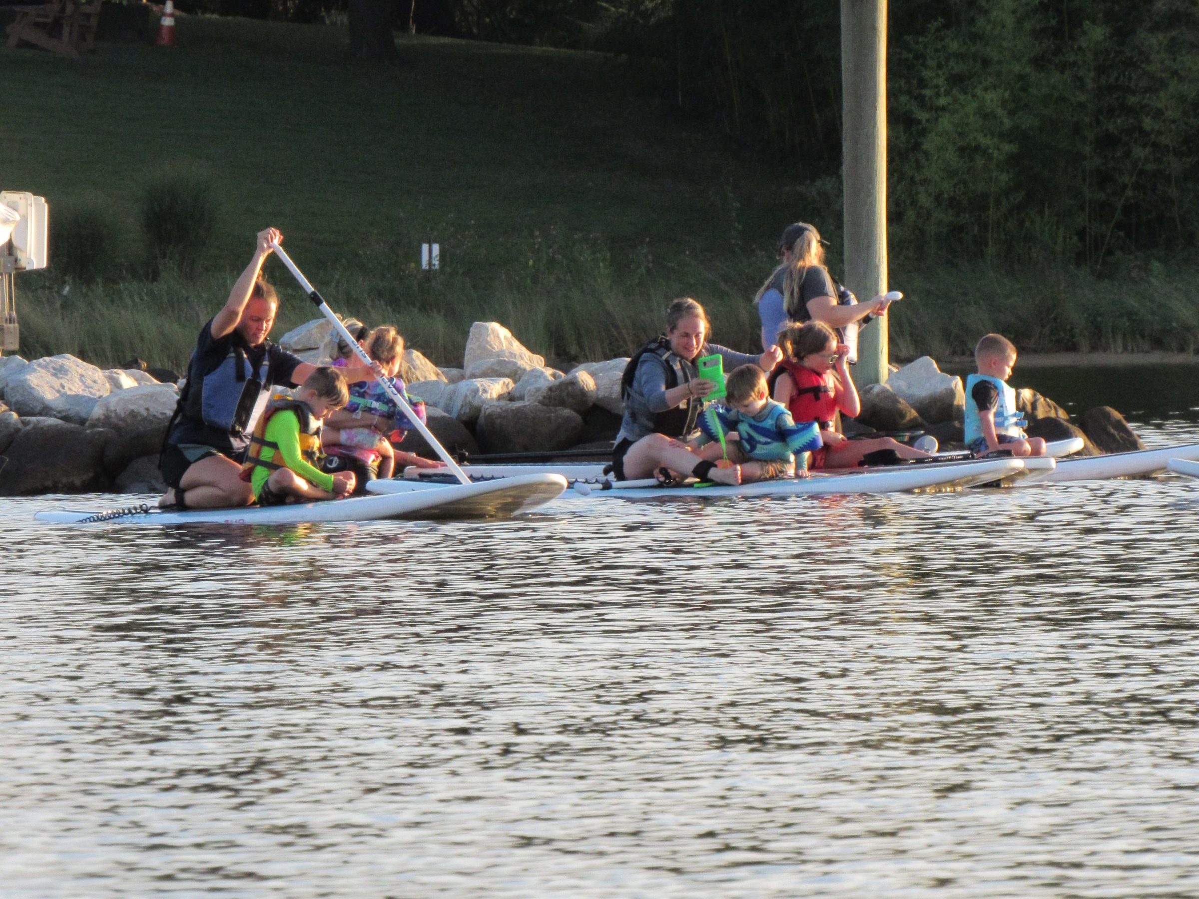 Two people paddleboarding with children on a calm waterway near a rocky shore.