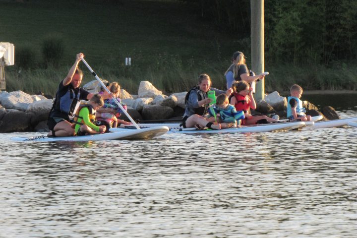 Two people paddleboarding with children on a calm waterway near a rocky shore.