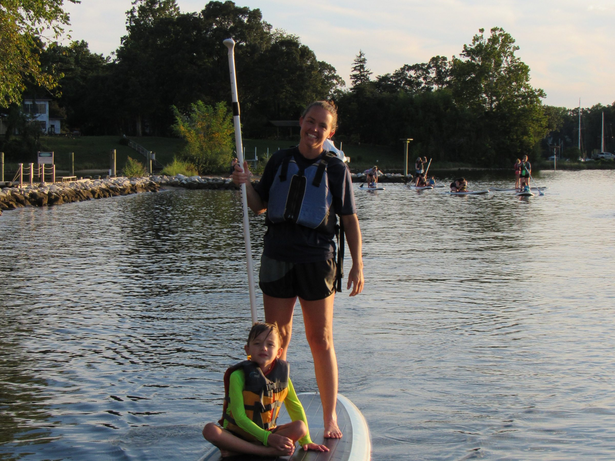 Two people on a stand-up paddleboard on a calm lake near a wooded shore.