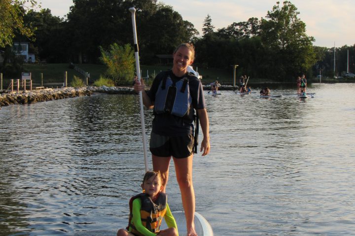 Two people on a stand-up paddleboard on a calm lake near a wooded shore.