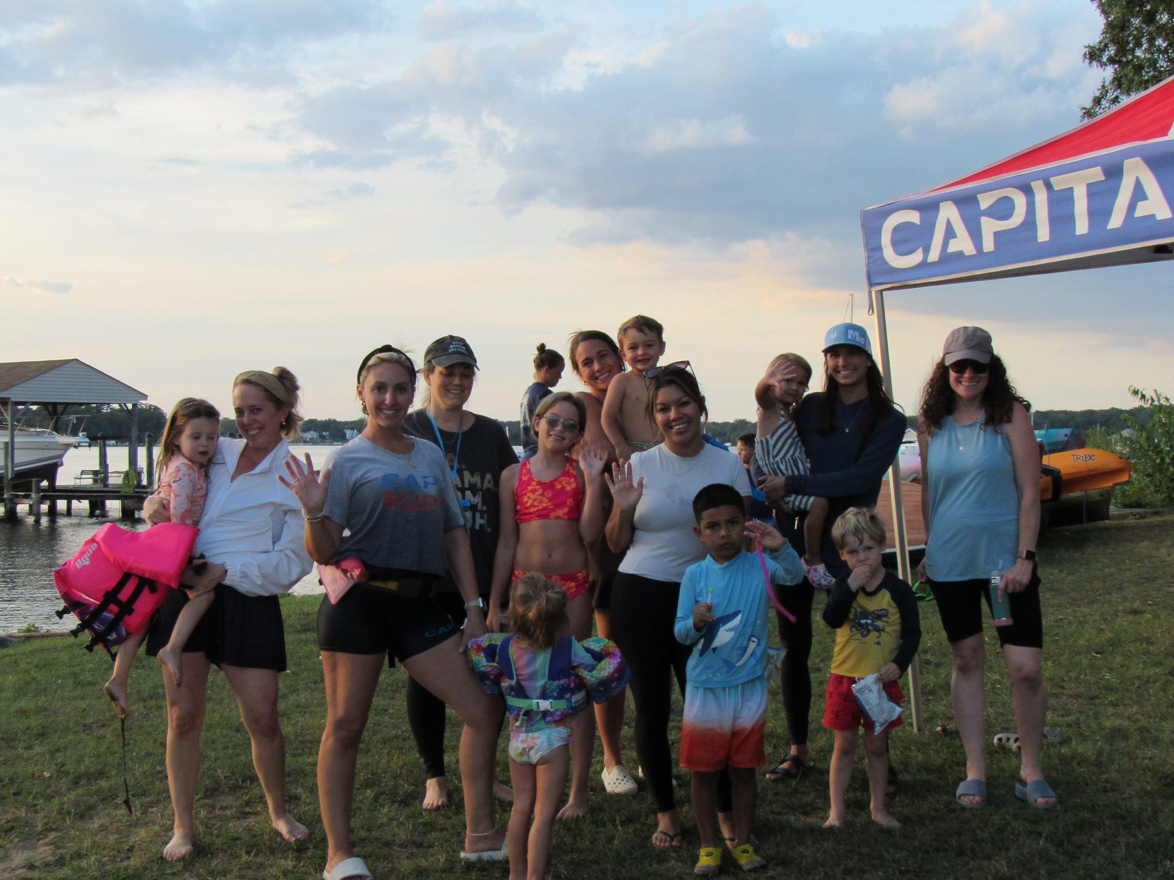 Group of adults and children posing by a lakeside with a colorful sky and tent in the background.
