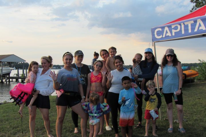 Group of adults and children posing by a lakeside with a colorful sky and tent in the background.