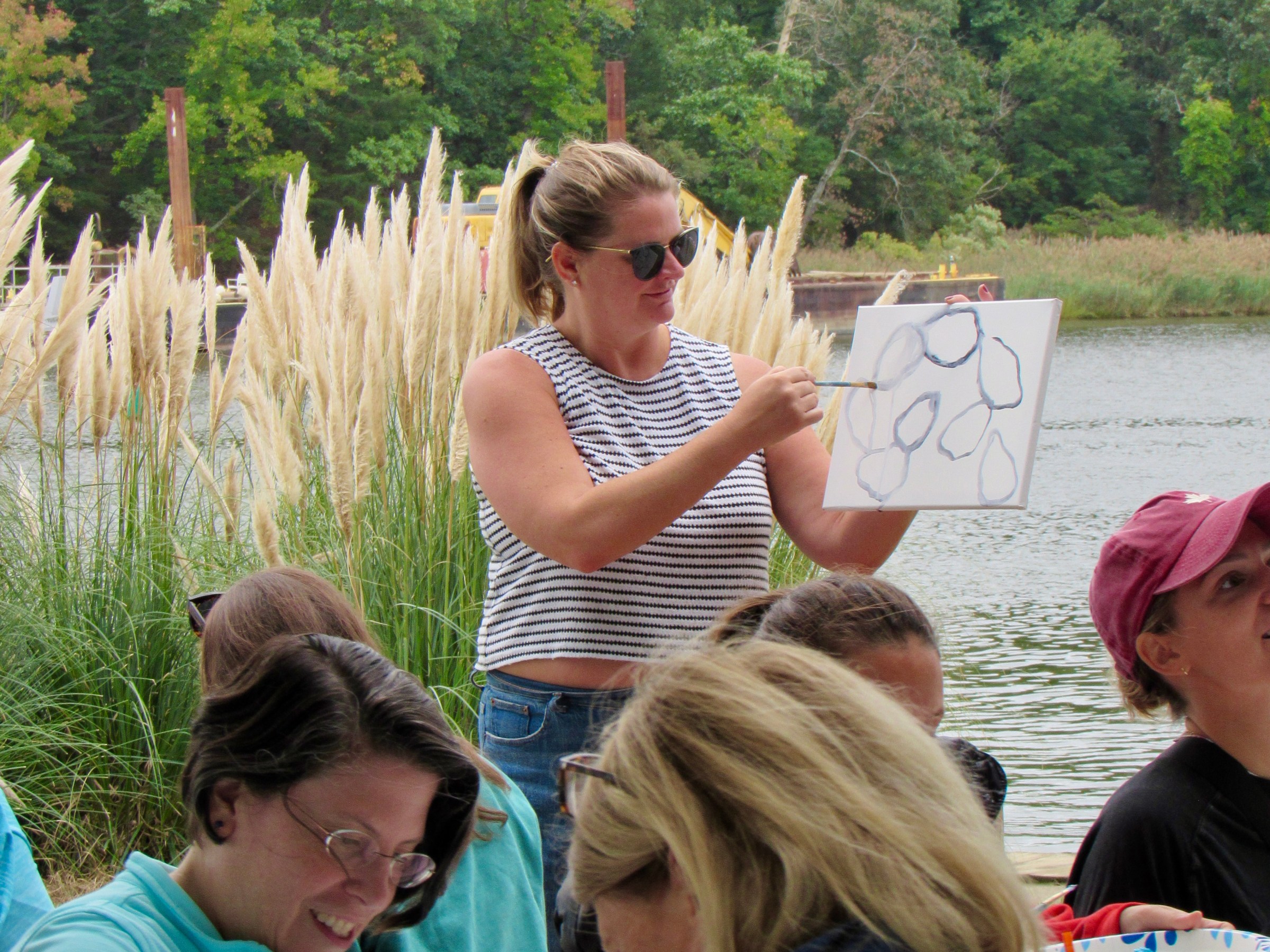 Woman holding painting at outdoor art class by a lake with participants seated, surrounded by greenery.