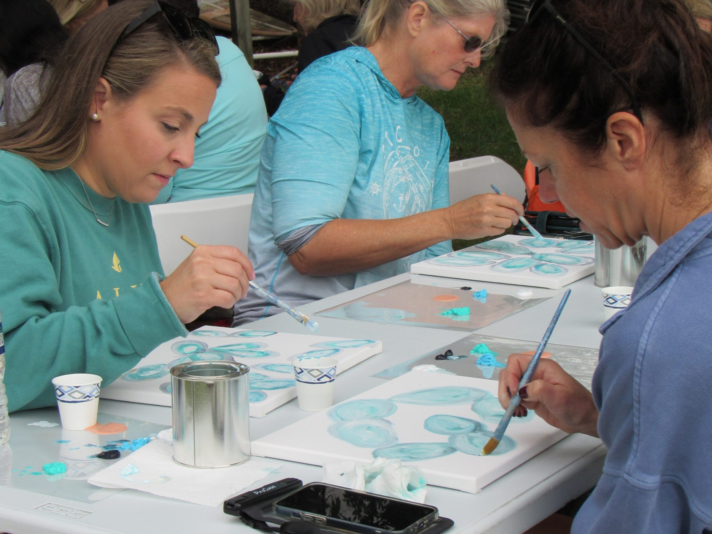 People painting at an outdoor table, focusing on canvas with blue paint.
