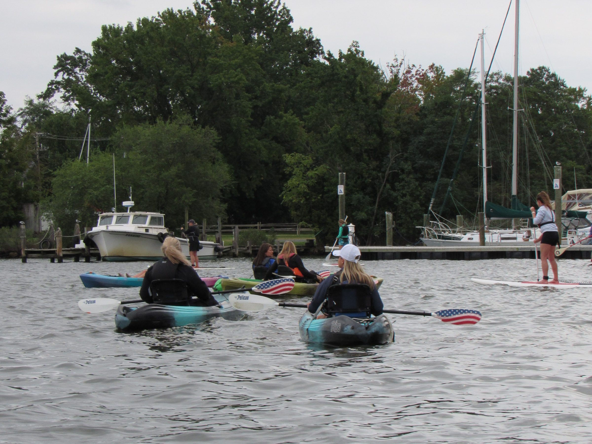 People kayaking with paddles decorated with U.S. flag, with boats and trees in the background.