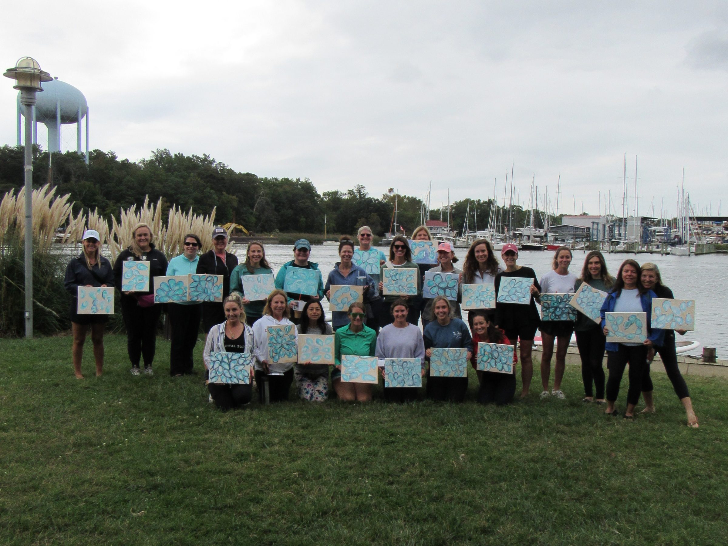 Group holding paintings outdoors by a waterfront under a cloudy sky.
