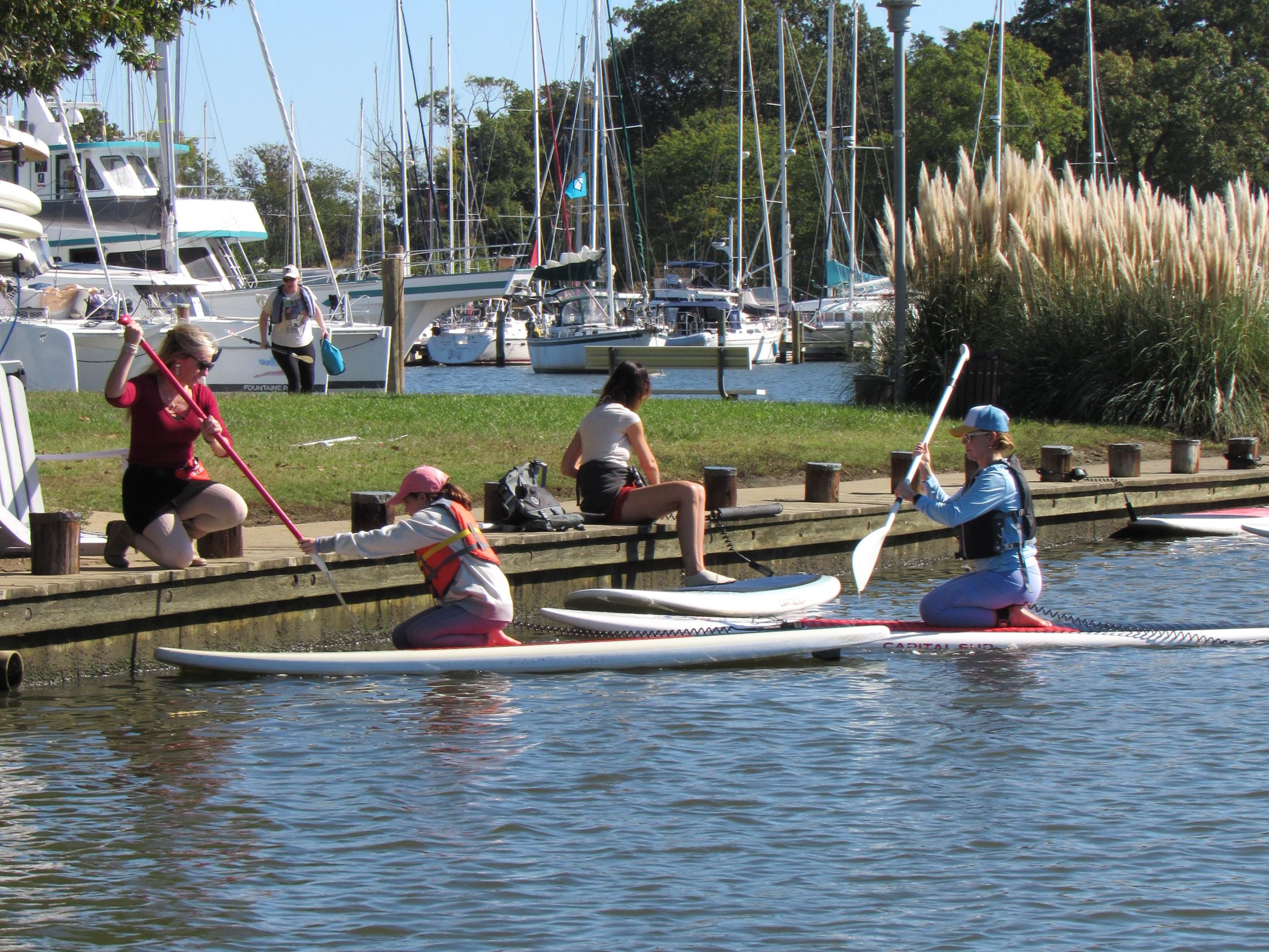 People on paddleboards and sitting by dock with sailboats in background.