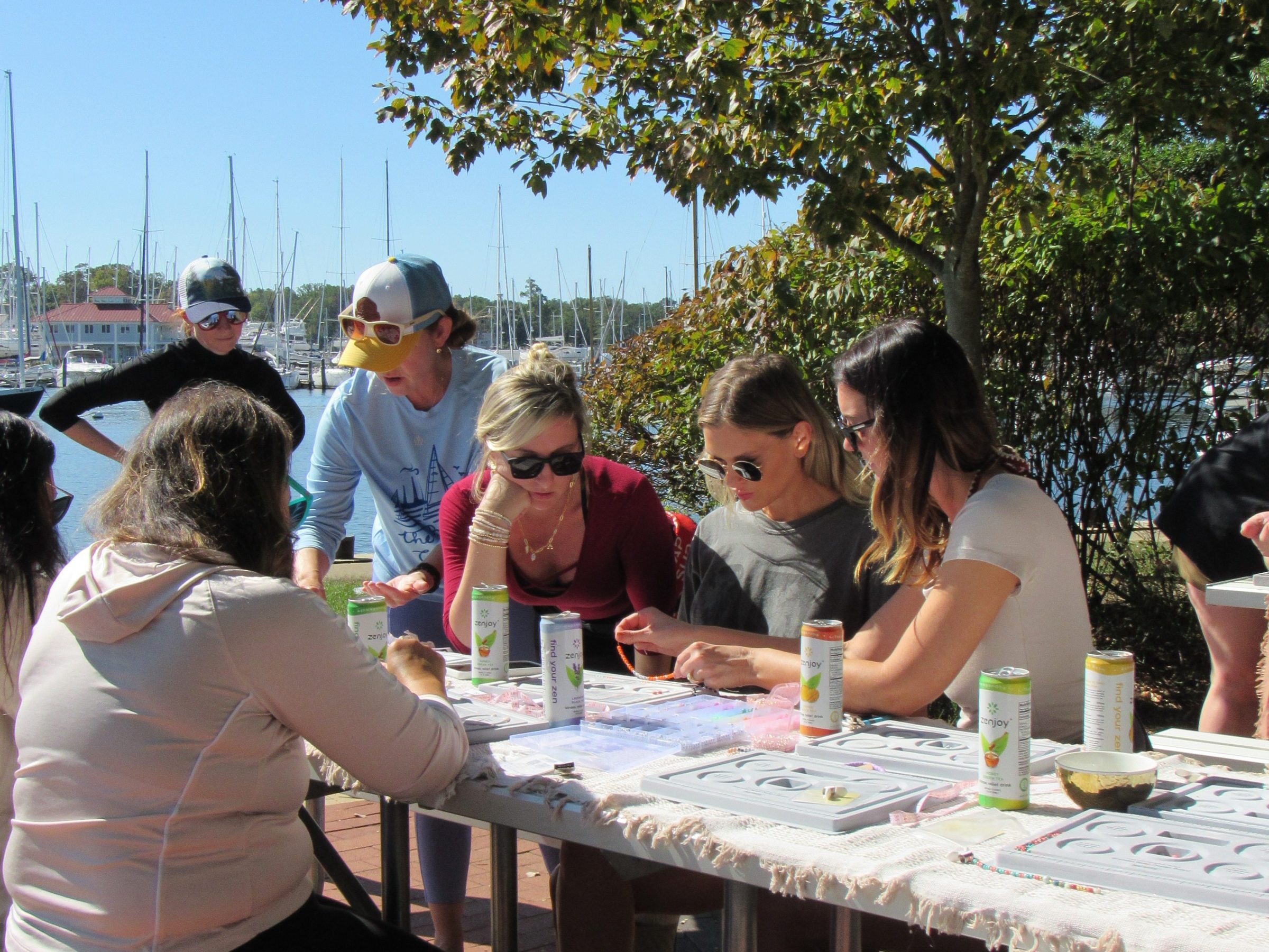 People crafting at outdoor table with supplies, under a tree by a marina.